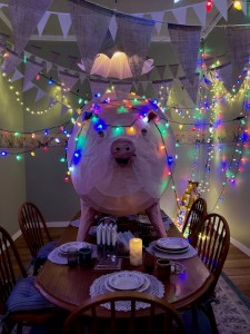 A room with a dining table set for a dinner party with fairy lights.  There is a very large replica pig standing in the middle of the table amongst the crockery
