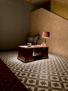 Theatre set showing office desk with light on a diamond motif carpet. There is a trussed up Zebra replica in one corner.